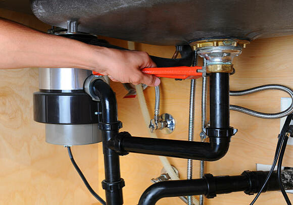 garbage-disposal-repair-sacramento Closeup of a plumber using a wrench to tighten a fitting beneath a kitchen sink. Only the man's hand and arm are visible. Horizontal format.